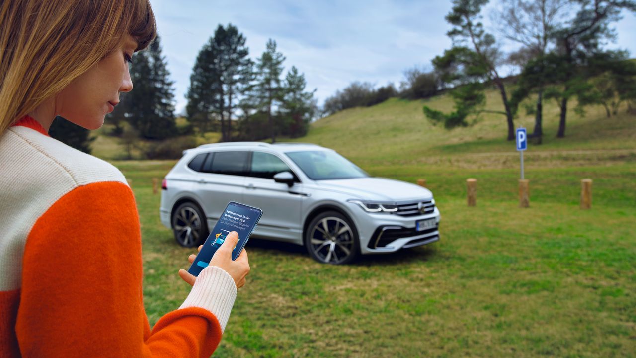 Eine Frau nutzt die Volkswagen App auf ihrem Smartphone, im Hintergrund steht der Tiguan Allspace auf einer Wiese