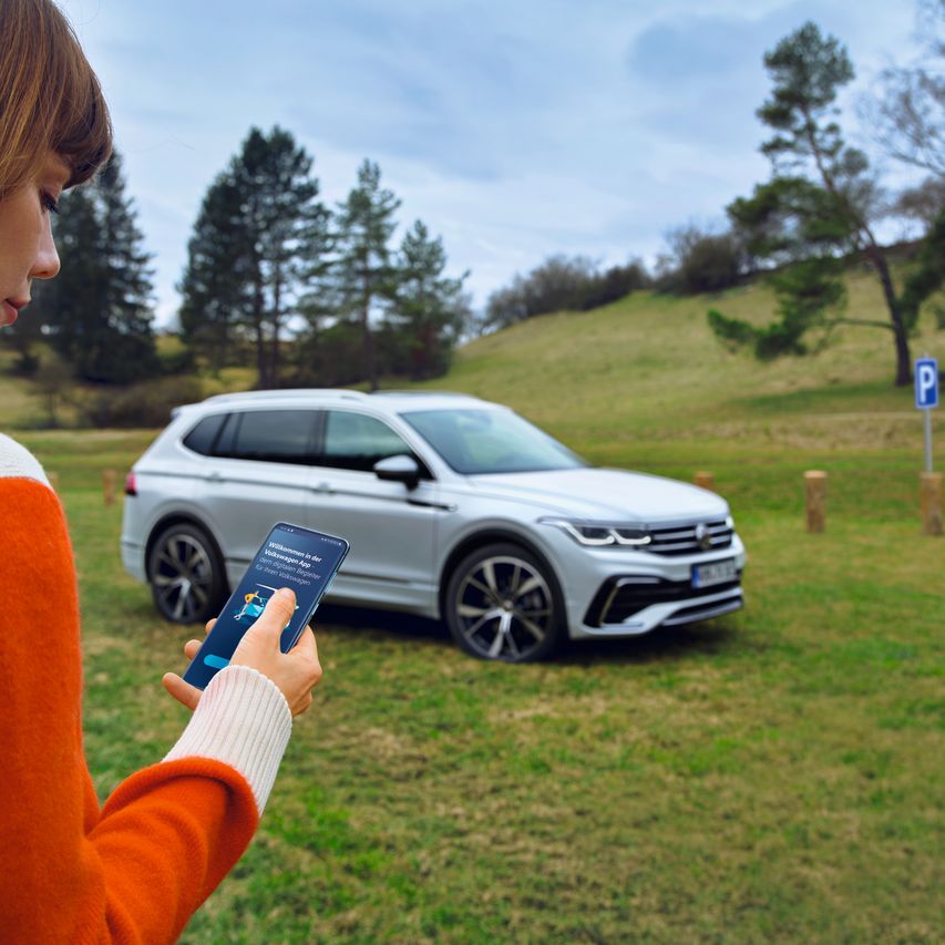Eine Frau nutzt die Volkswagen App auf ihrem Smartphone, im Hintergrund steht der Tiguan Allspace auf einer Wiese