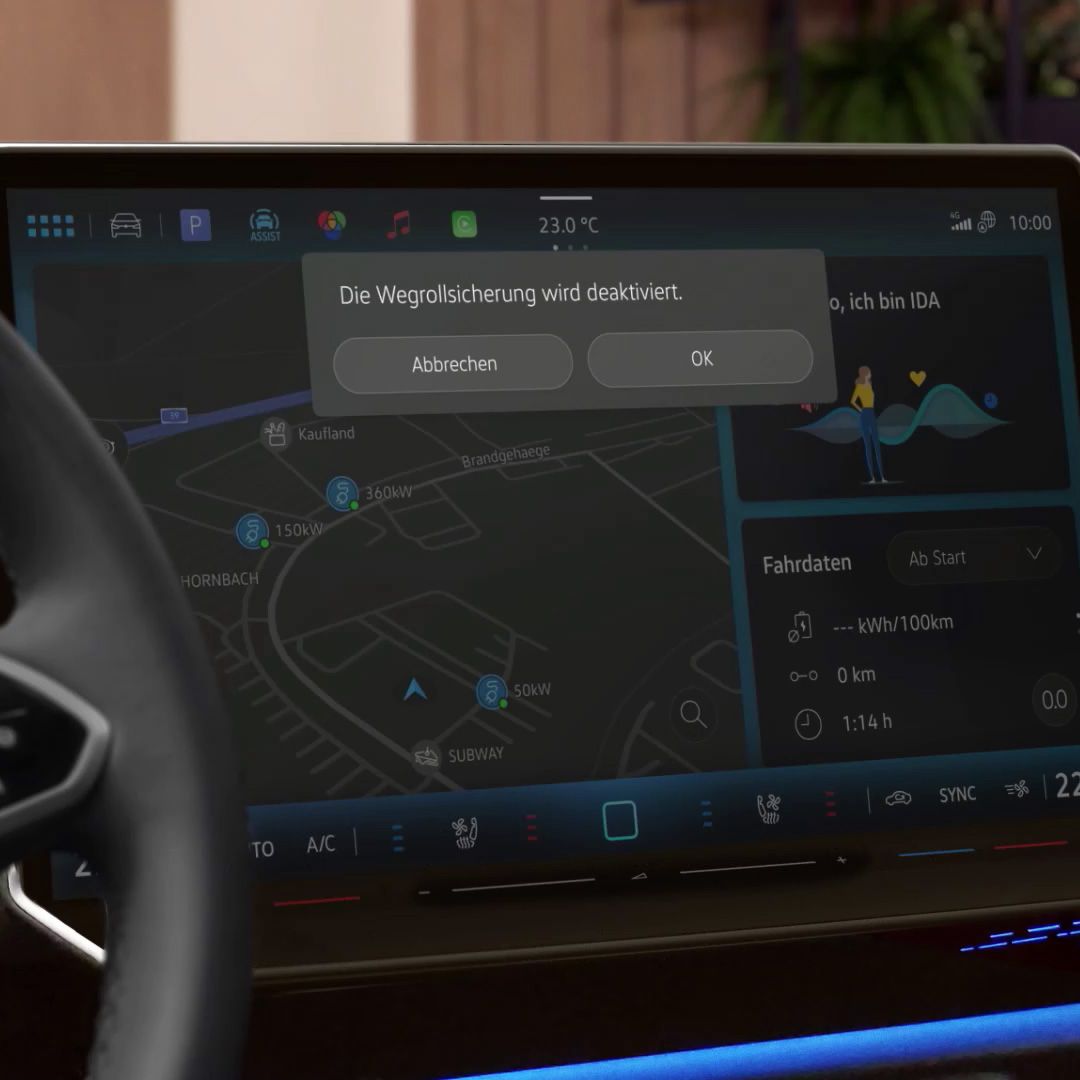 Ein Infotainment-Bildschirm im Auto zeigt die Nachricht "Die Wegrollsicherung wird deaktiviert" mit den Optionen "Abbrechen" und "OK". Im Hintergrund sind Navigations- und Informationsanzeigen sichtbar. Ein Teil des Lenkrads ist ebenfalls zu sehen.