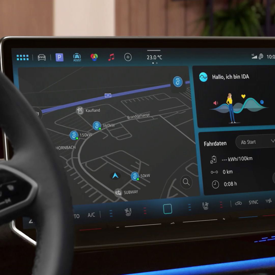Nahaufnahme eines digitalen Navigationsbildschirms in einem ID.7 Tourer mit einer Karte, die verschiedene Ladestationen zeigt. Rechts im Bild informiert eine virtuelle Assistentin namens IDA den Fahrer über den Status und die Fahrdaten. Das Steuer des Fahrzeugs ist ebenfalls teilweise sichtbar.