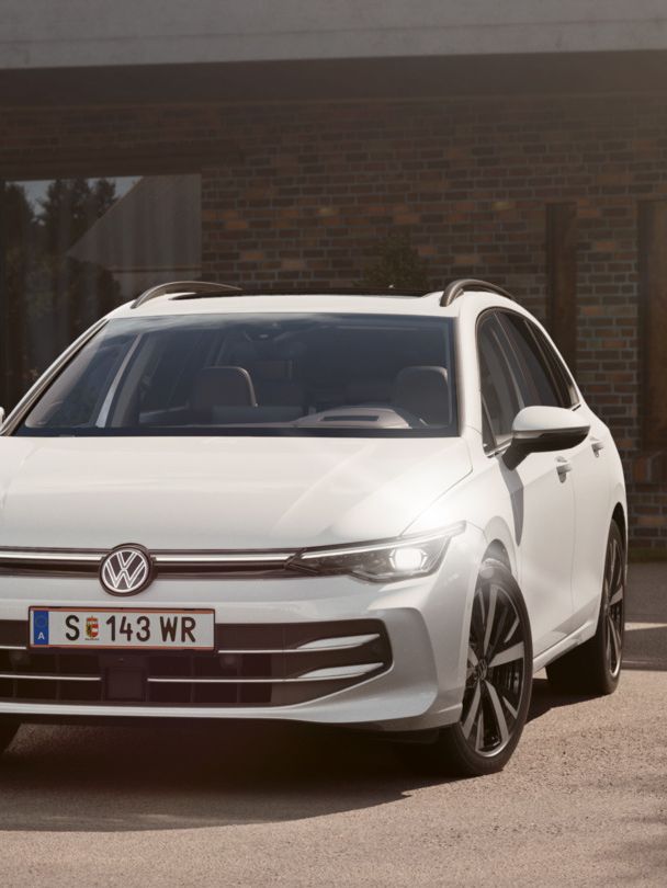 Ein weißer VW Golf Variant parkt in der Auffahrt eines Bungalows mit aktivem Tagfahrlicht.