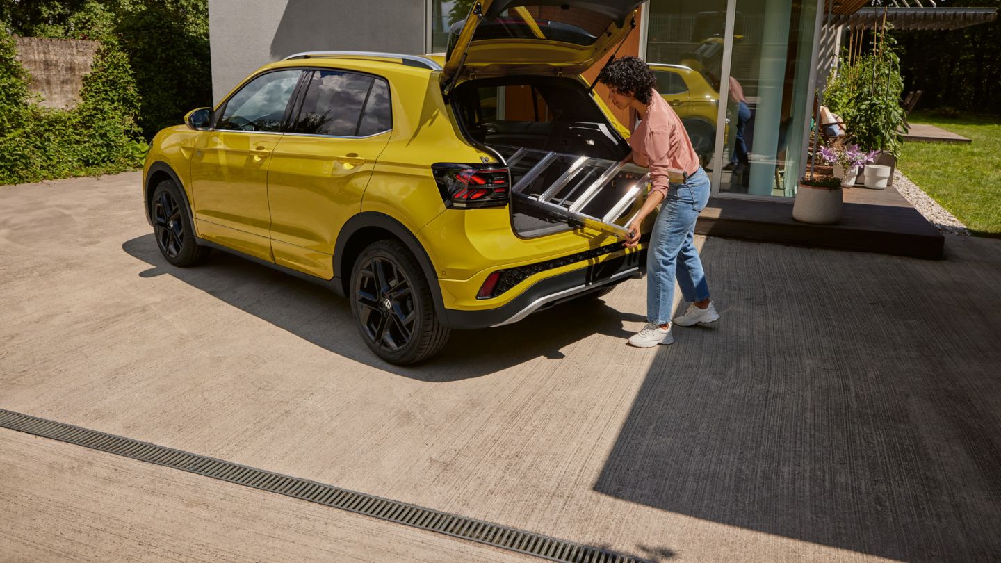 Eine junge Frau schiebt eine Leiter in den Kofferraum eines gelben VW T-Cross.