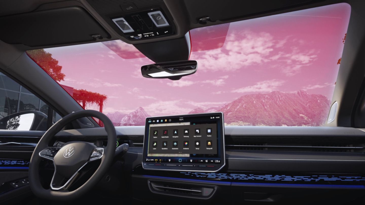 Innenraum eines modernen Autos mit großer digitaler Anzeige in der Mittelkonsole. Das Lenkrad und die Frontscheibe sind sichtbar, durch die man eine Landschaft mit Bergen und einem roten Himmel sieht.