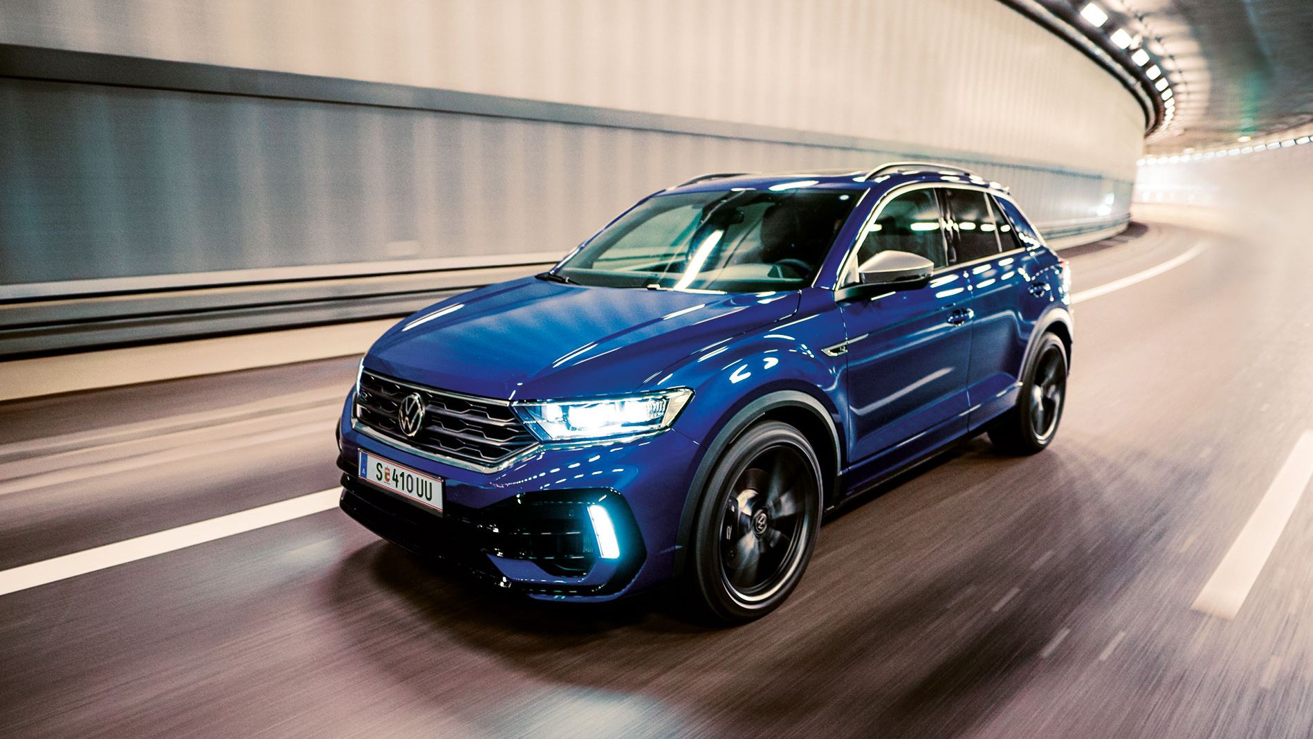 Ein blauer VW T-Roc fährt schnell durch einen beleuchteten Tunnel. Das Auto sieht modern und sportlich aus. Die Straßenmarkierungen sind deutlich auf dem Boden sichtbar.