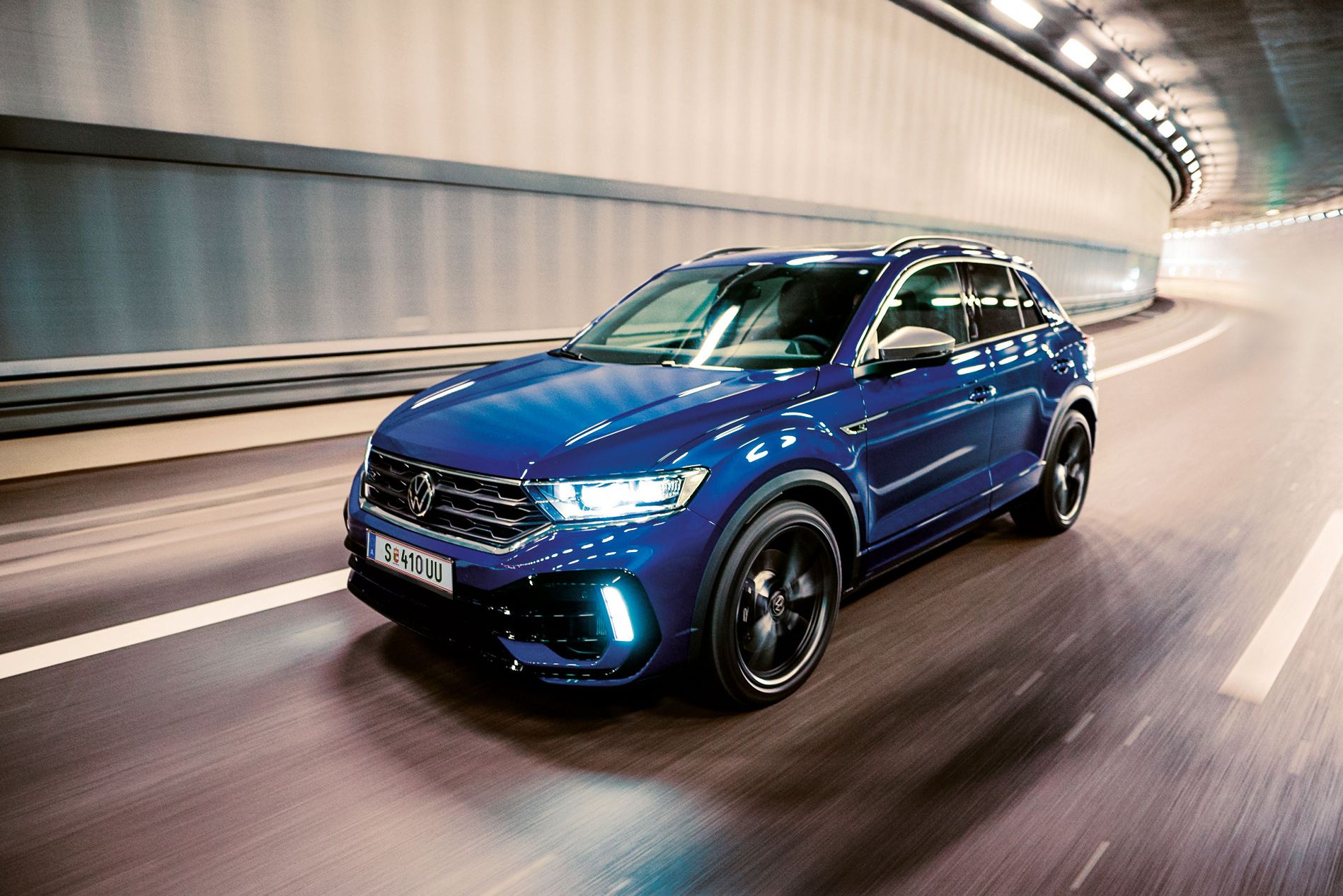 Ein blauer VW T-Roc fährt schnell durch einen beleuchteten Tunnel. Das Auto sieht modern und sportlich aus. Die Straßenmarkierungen sind deutlich auf dem Boden sichtbar.