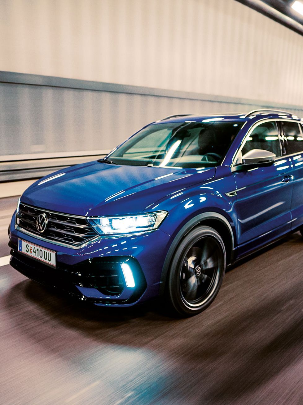 Ein blauer VW T-Roc fährt schnell durch einen beleuchteten Tunnel. Das Auto sieht modern und sportlich aus. Die Straßenmarkierungen sind deutlich auf dem Boden sichtbar.