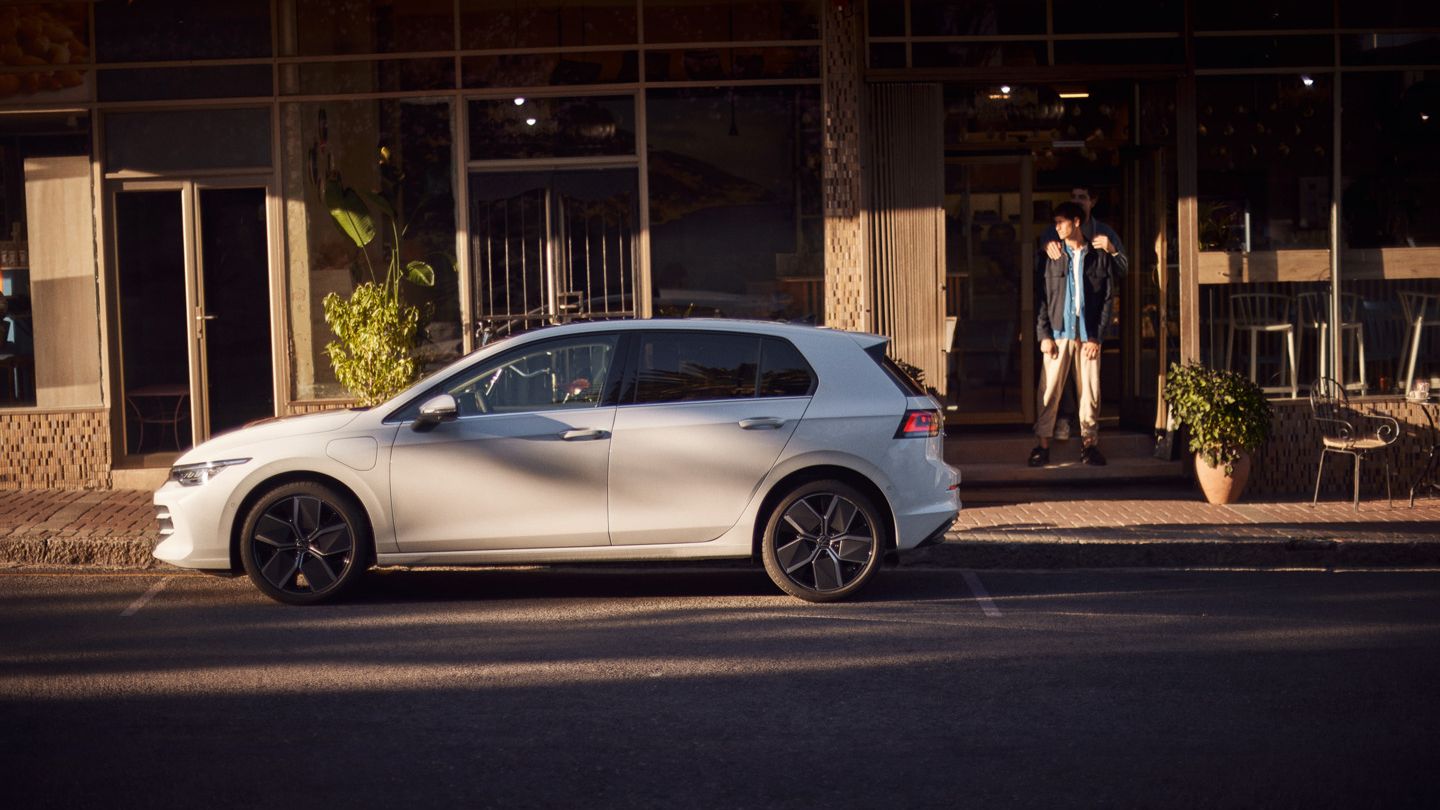 Ein weißer Golf eHybrid parkt am Straßenrand vor einem Café. 