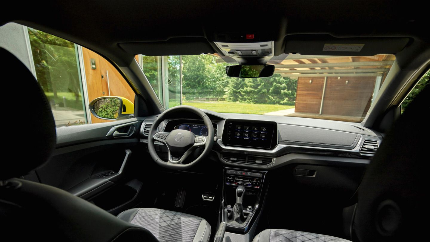 Blick vom Rücksitz aus ins Cockpit eines VW T-Cross R-Line und durch die Frontscheibe auf ein Haus mit Garten.