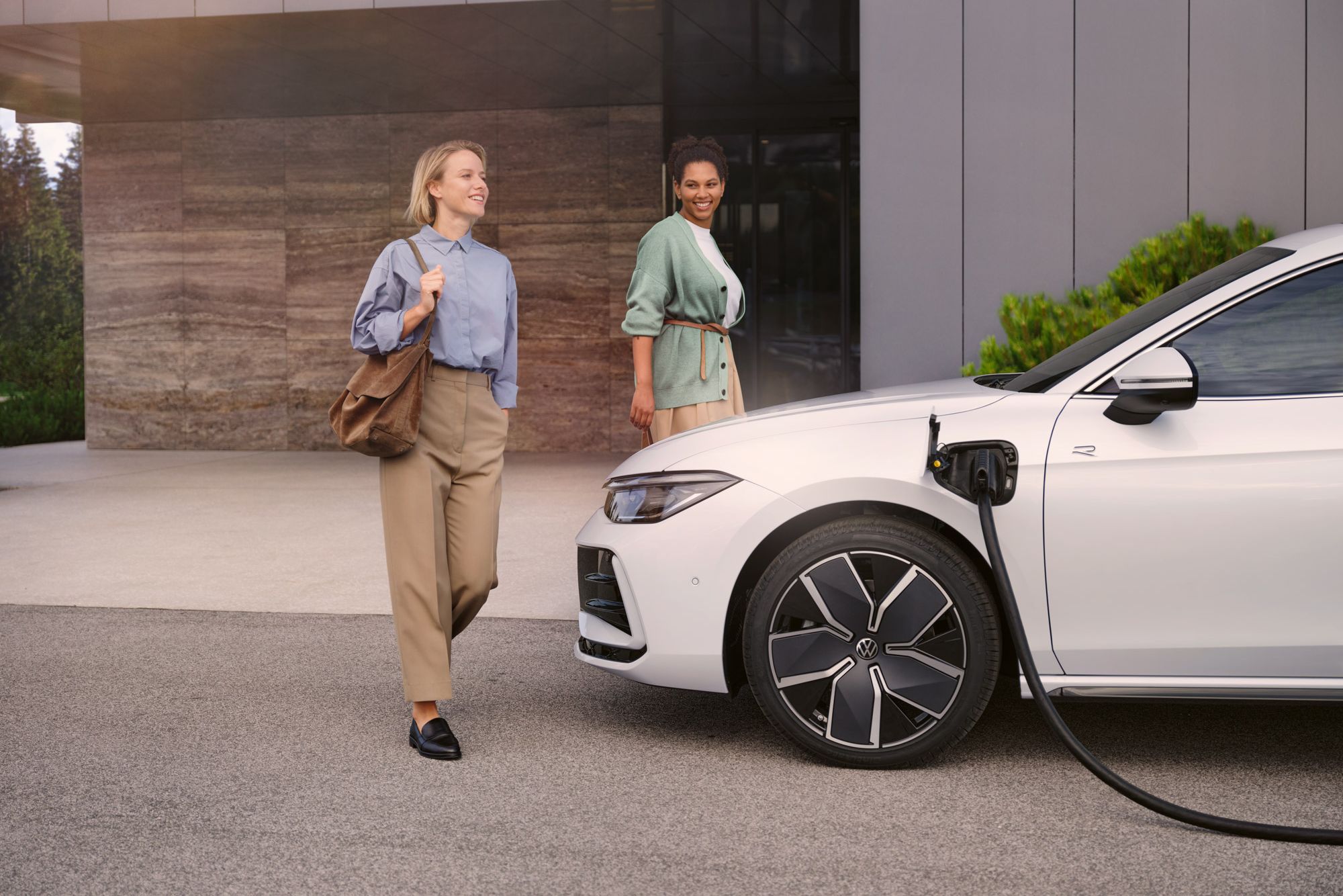 Weißer VW Passat steht mit eingestecktem Ladekabel vor einem Haus, zwei Frauen gehen auf den Wagen zu.