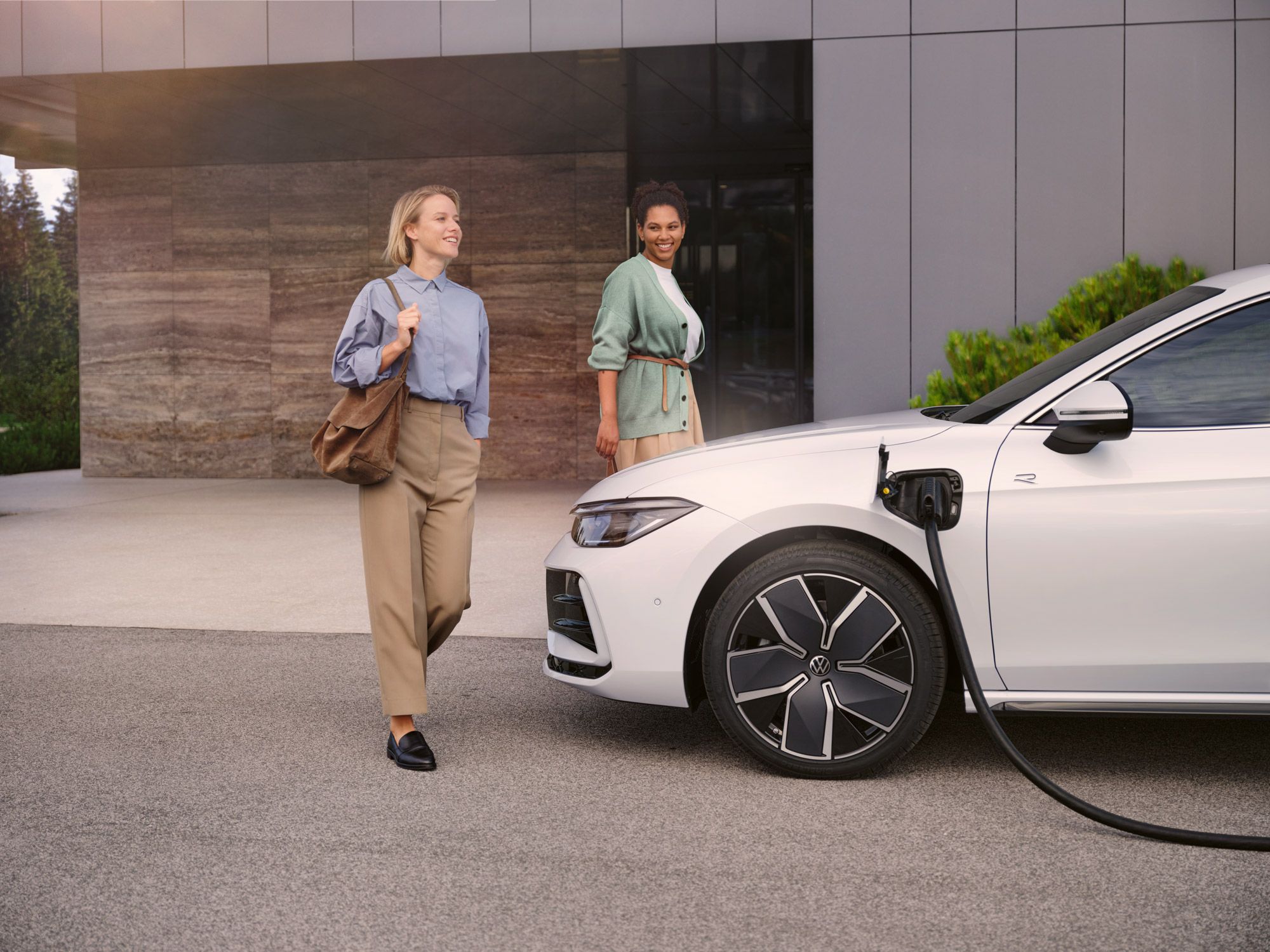 Weißer VW Passat steht mit eingestecktem Ladekabel vor einem Haus, zwei Frauen gehen auf den Wagen zu.