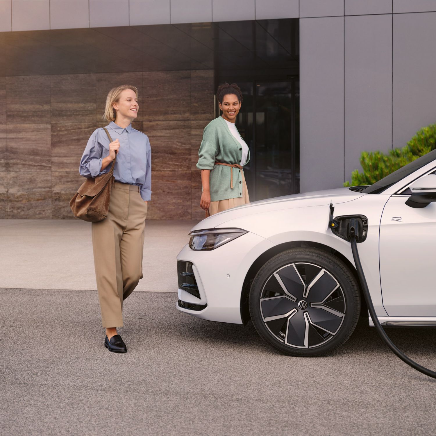 Weißer VW Passat steht mit eingestecktem Ladekabel vor einem Haus, zwei Frauen gehen auf den Wagen zu.