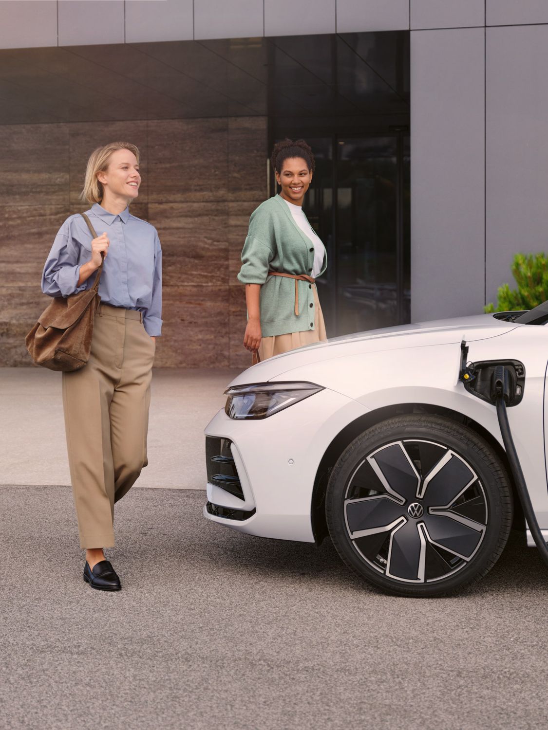 Weißer VW Passat steht mit eingestecktem Ladekabel vor einem Haus, zwei Frauen gehen auf den Wagen zu.