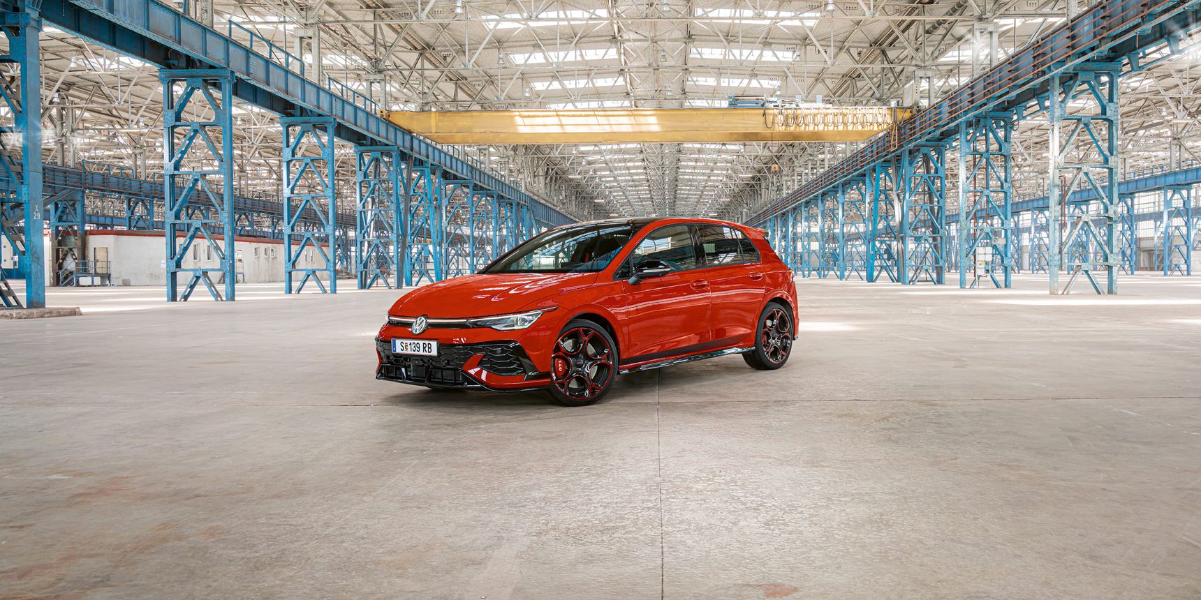 Ein roter Golf GTI steht in einer großen, leeren Industriehalle mit blauen Stahlträgern. Das Dach der Halle ist aus Glas und lässt viel Licht herein.