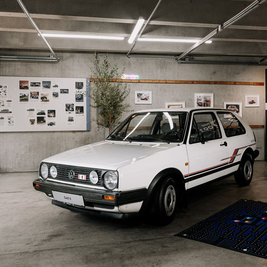 Ein weißer VW Golf 2 GTI steht in einer Garage mit dekorativen Bildern an der Wand. Das Auto ist gut erhalten und sauber. Am Boden liegt eine großformatige Pac-Man-Matte.