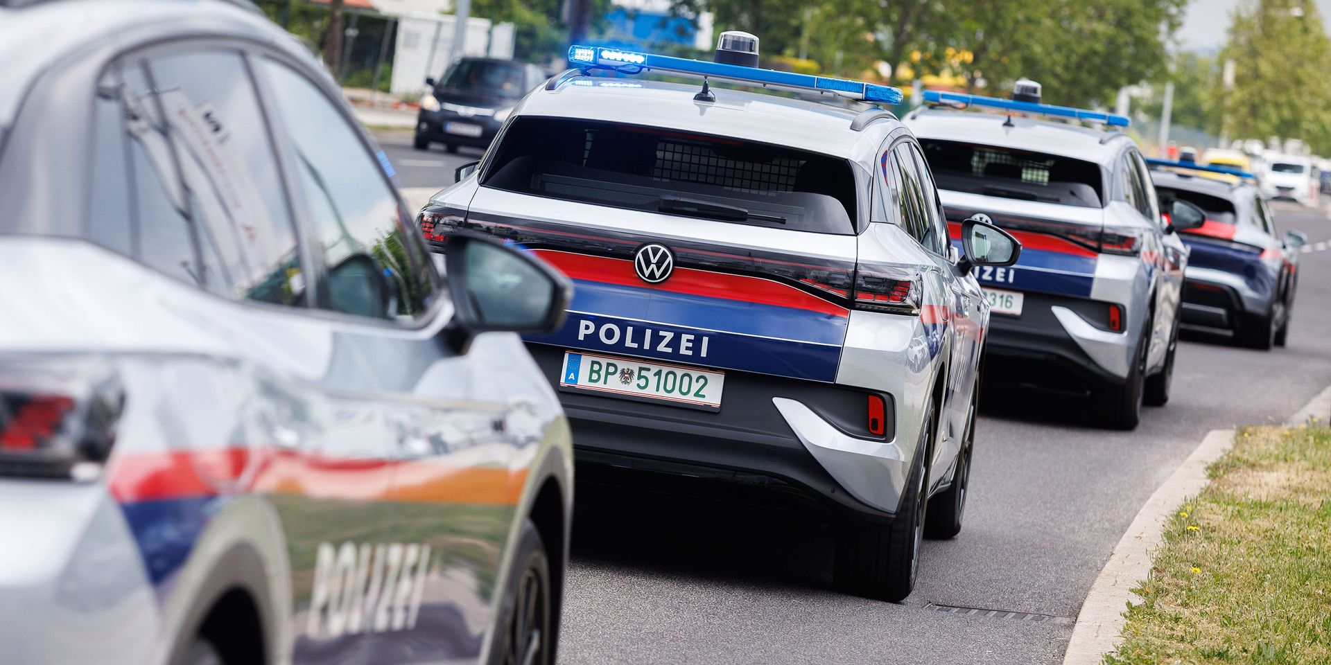 Eine Reihe von österreichischen Polizeiautos fährt auf einer Straße. Die Fahrzeuge haben Blaulichter und sind weiß mit blauen und roten Streifen. Im Hintergrund sind Bäume und Gebäude zu sehen.