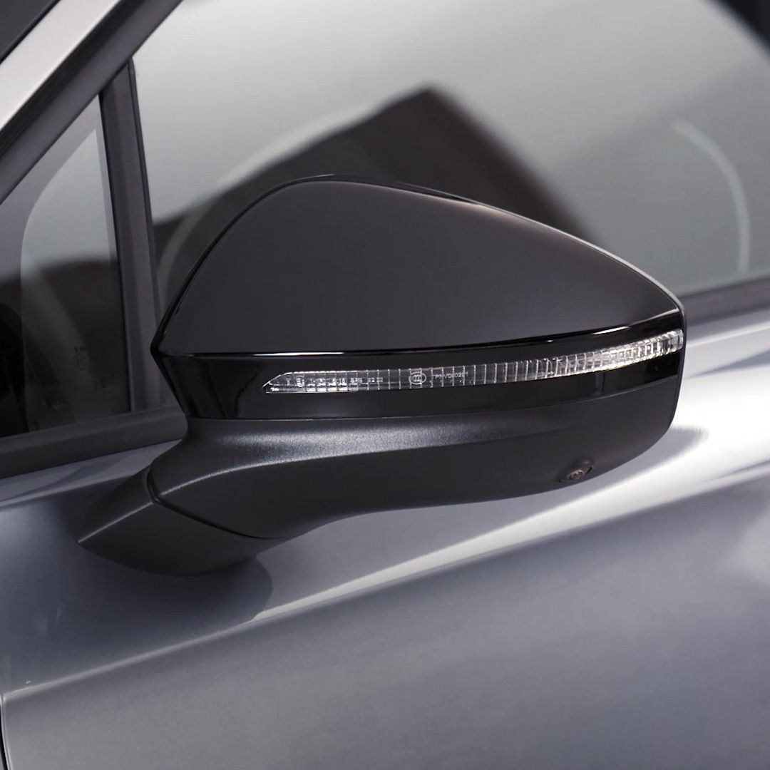 Nahaufnahme eines schwarzen Seitenspiegels an einem silbernen Auto. Der Spiegel hat eine eingebaute LED-Leuchte zum Blinken. Im Hintergrund ist ein Teil des Fensters und der Tür des Autos zu sehen.