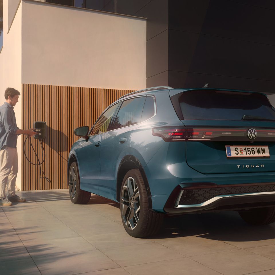 Blauer VW Tiguan eHybrid steht vor einem modernen Wohnhaus. Ein Mann daneben hält den Stecker einer Ladestation in der Hand.
