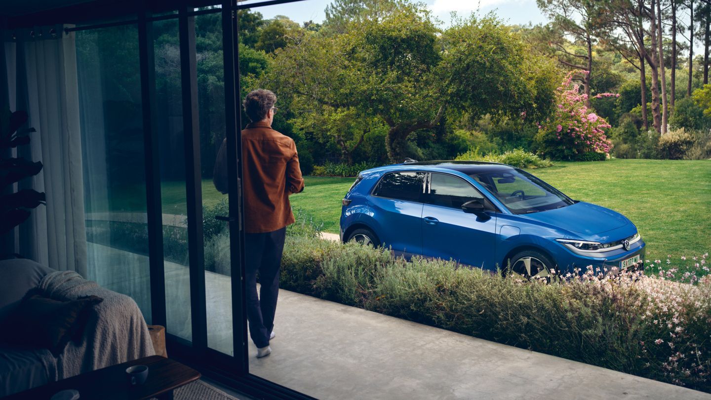 Blick durch eine offene Terrassentür in einen grünen Garten, in der Auffahrt steht ein blauer VW ID. Polo Style.