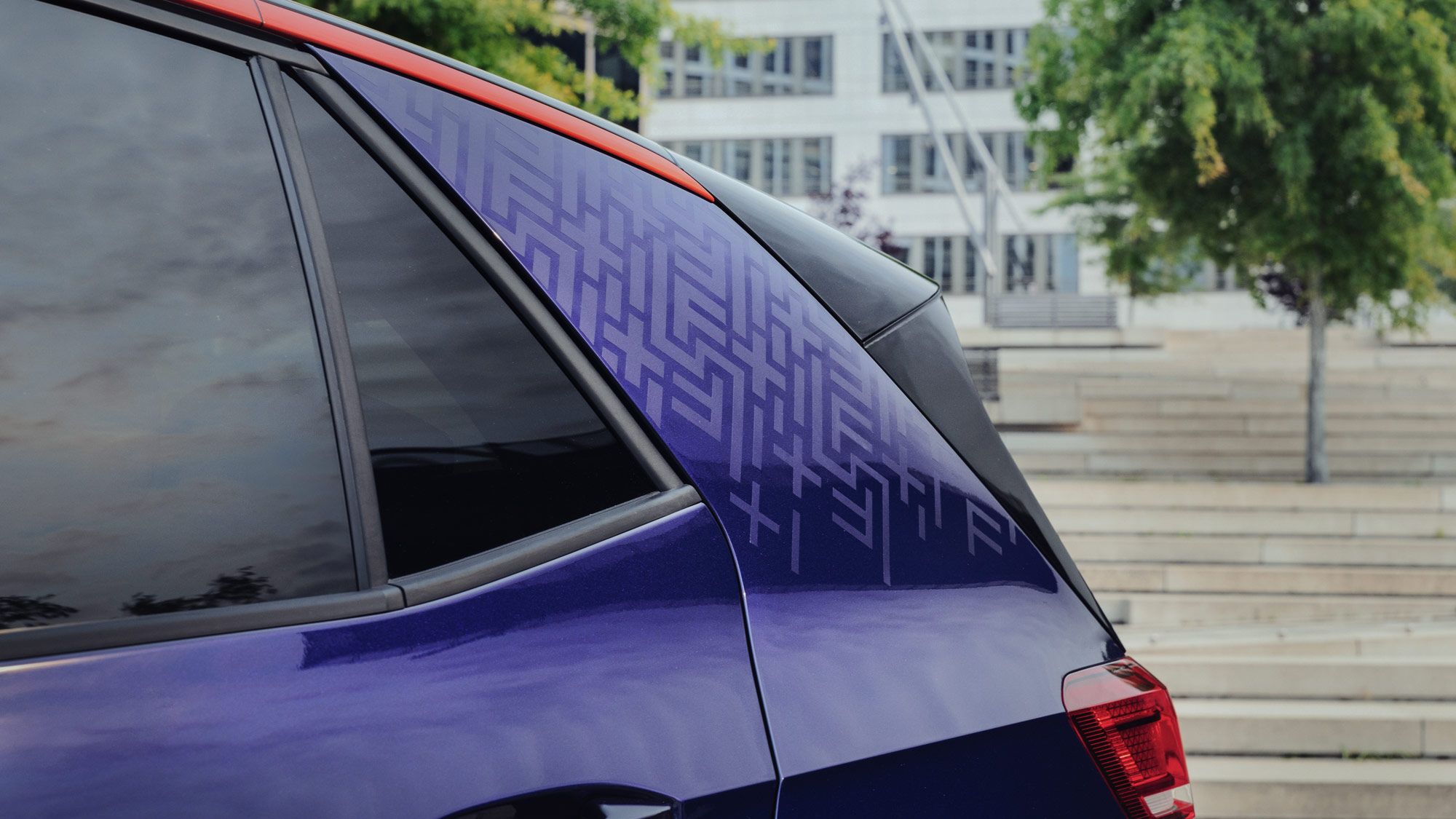 Rückansicht eines dunkelblauen Autos mit auffälliger rot-schwarzer Kontrastierung vom Dach zum hinteren Fensterbereich. Das Auto steht im Freien vor einem modernen, weißen Gebäudekomplex. Die Rücklehne eines Sitzplatztreppenbereichs ist ebenfalls erkennbar im Hintergrund.