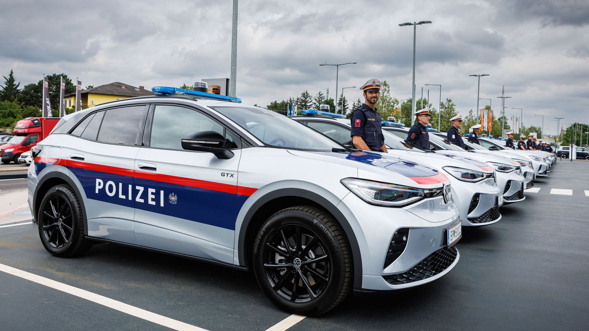 Eine Reihe von Polizeifahrzeugen ist geparkt, es sind silberne ID.4 GTX mit blauen und roten Streifen. Im Vordergrund steht ein Polizist neben einem Fahrzeug. Der Himmel ist bewölkt.