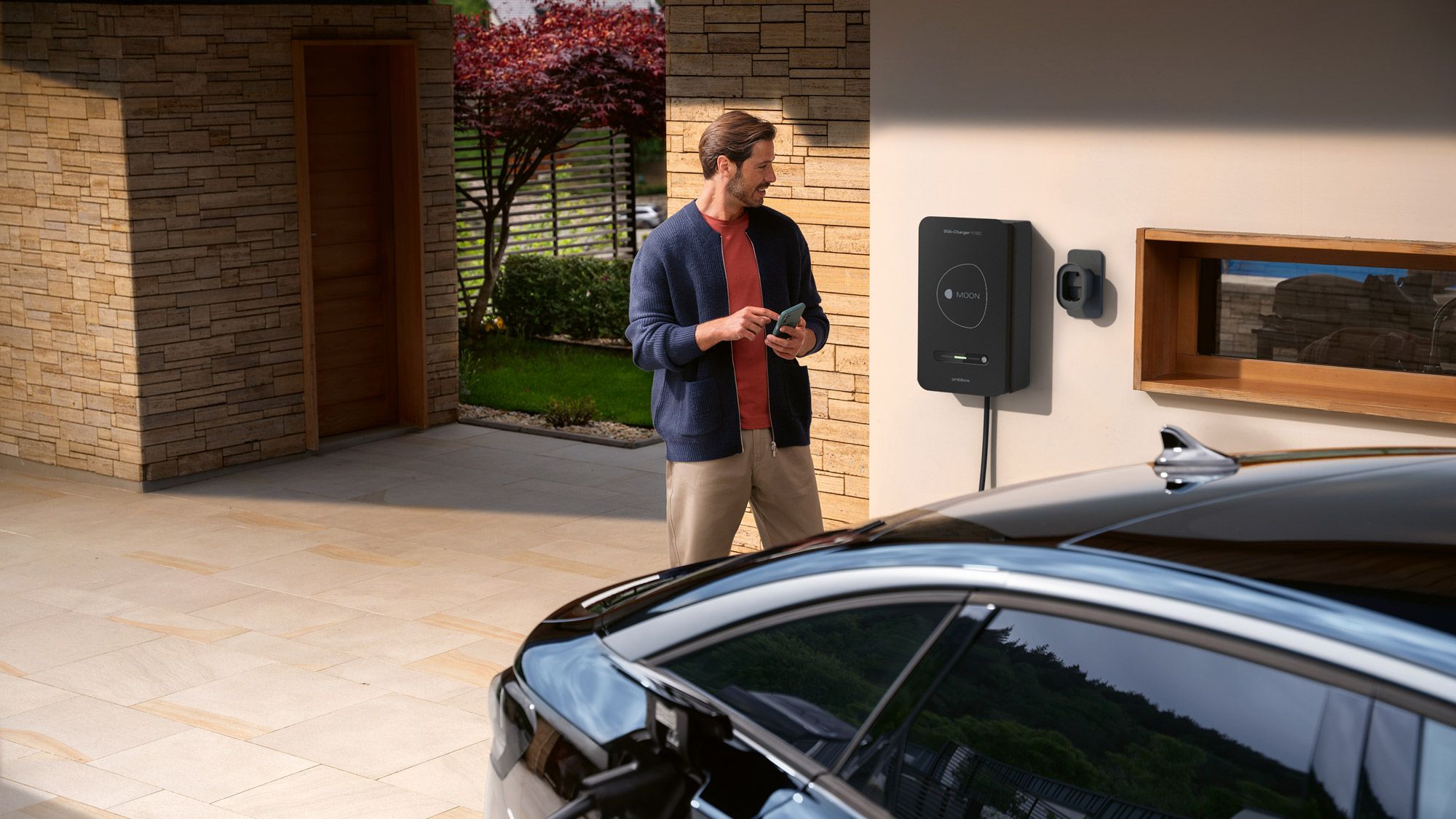 Ein Mann steht in einer modernen Einfahrt neben einem Haus und bedient eine an der Wand montierte Ladestation. Im Vordergrund ist ein schwarzes Elektroauto zu sehen, das über ein Kabel mit der Wallbox verbunden ist. Die Szene wirkt gepflegt und hochwertig, mit Steinfassade, hellen Bodenplatten und einem Baum im Hintergrund.