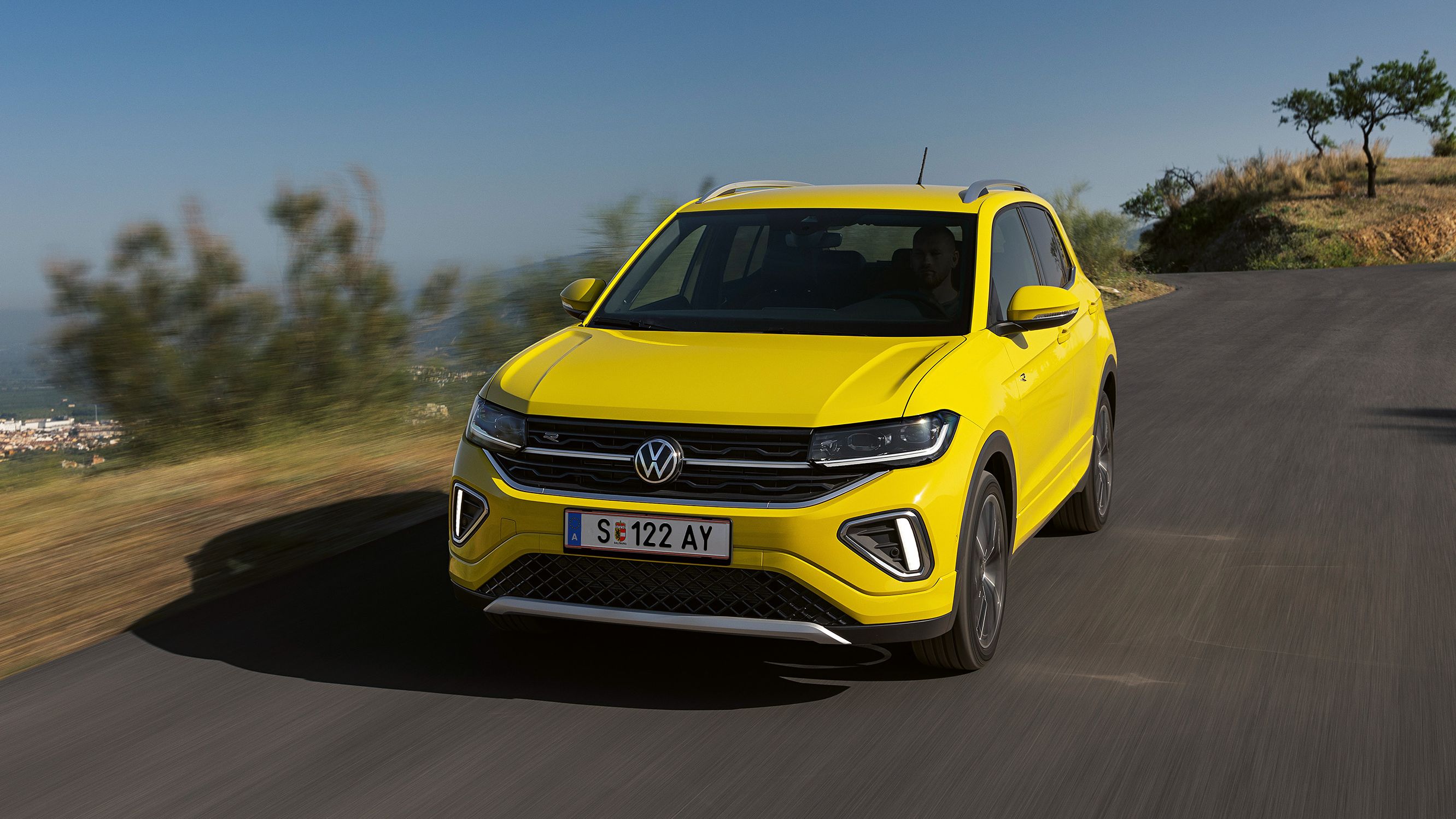 Gelber Volkswagen SUV fährt auf einer Straße in einer ländlichen Gegend. Hintergrund zeigt verschwommene Natur und stark bewölkten Himmel. Das Fahrzeug ist frontal zu sehen.