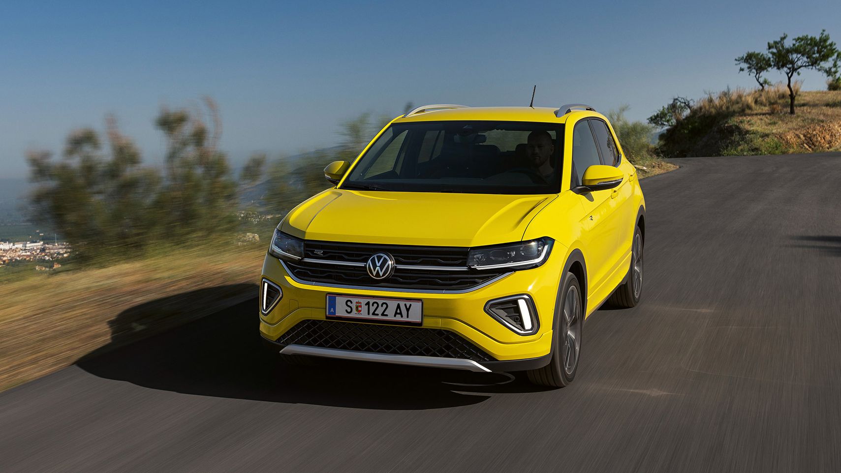 Gelber Volkswagen SUV fährt auf einer Straße in einer ländlichen Gegend. Hintergrund zeigt verschwommene Natur und stark bewölkten Himmel. Das Fahrzeug ist frontal zu sehen.