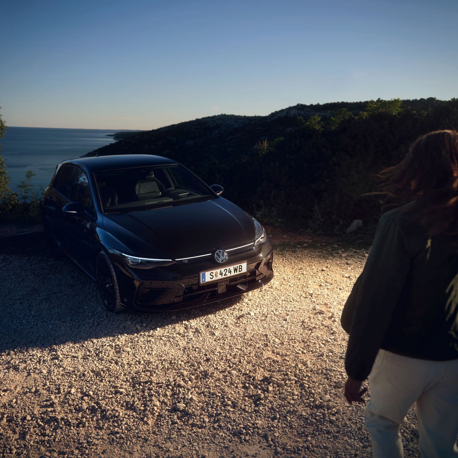Eine Frau geht auf einen schwarzen VW Golf R zu, das auf einem Kiesweg vor einer Küstenlandschaft geparkt ist. Die Sonne geht unter und taucht die Szene in ein warmes Licht. Im Hintergrund ist das Meer und einige Bäume zu sehen.