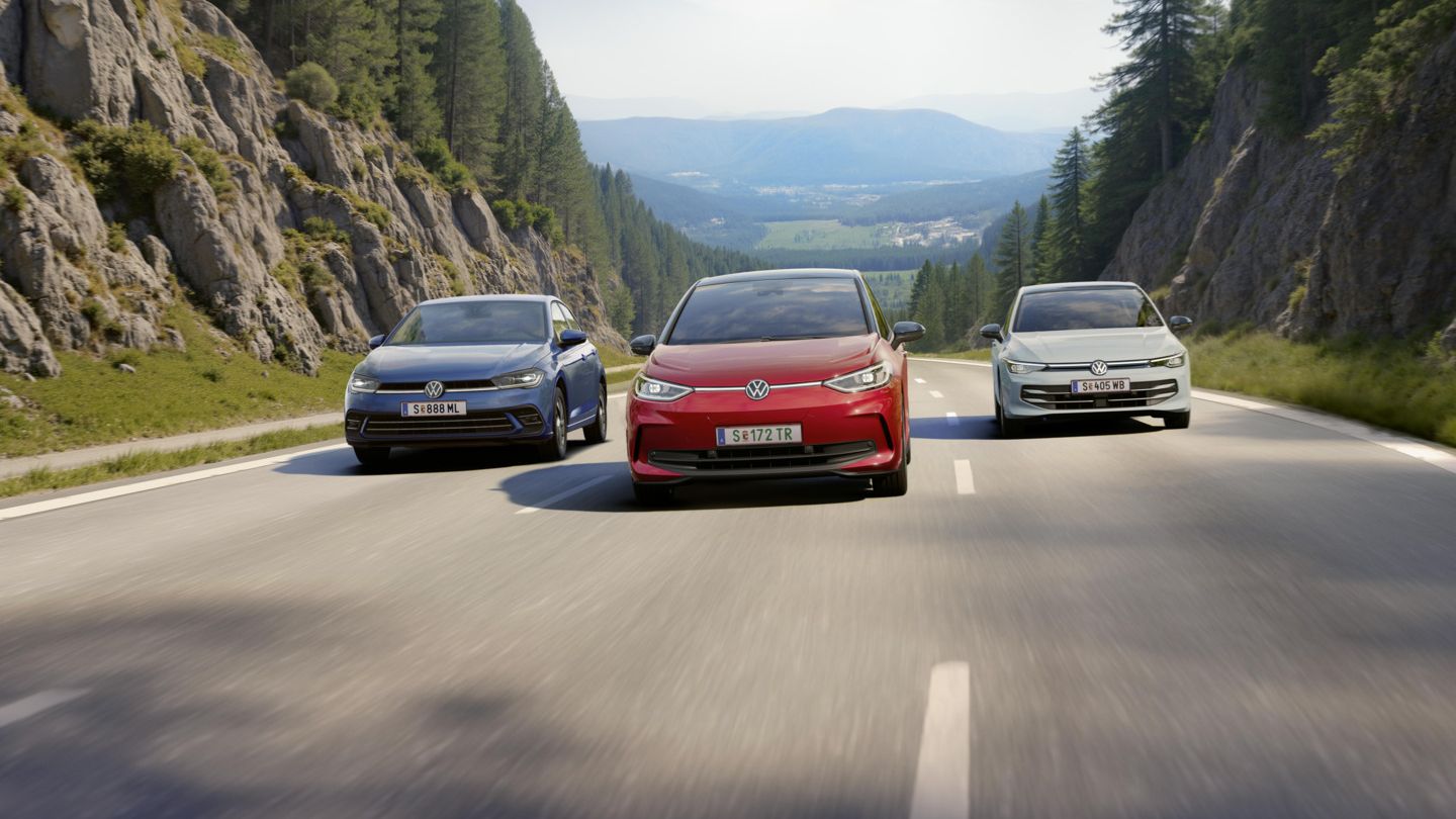Drei VW fahren nebeneinander über eine dreispurige Landstraße. auf der mittleren Spur fährt ein roter VW ID.3, links daneben ein weißer Golf und rechts ein blauer Polo. Die Straße führt durch eine Waldlandschaft und ist umgeben von Felswänden und die Sonne scheint. 