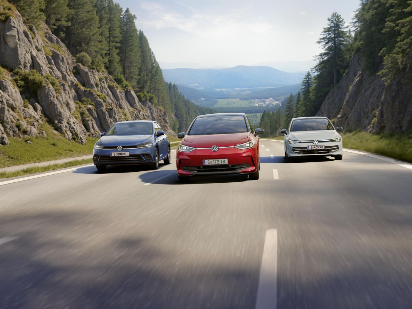 Drei VW fahren nebeneinander über eine dreispurige Landstraße. auf der mittleren Spur fährt ein roter VW ID.3, links daneben ein weißer Golf und rechts ein blauer Polo. Die Straße führt durch eine Waldlandschaft und ist umgeben von Felswänden und die Sonne scheint. 
