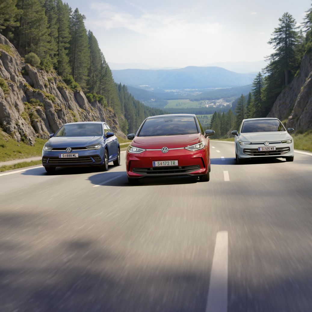 Drei VW fahren nebeneinander über eine dreispurige Landstraße. auf der mittleren Spur fährt ein roter VW ID.3, links daneben ein weißer Golf und rechts ein blauer Polo. Die Straße führt durch eine Waldlandschaft und ist umgeben von Felswänden und die Sonne scheint. 