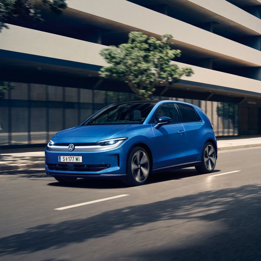 Ein blauer VW ID. Polo Style fährt durch eine Stadt mit grünen Bäumen am Straßenrand.