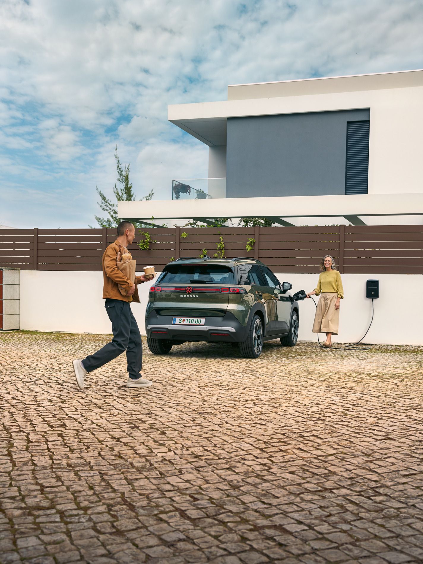 Eine Frau steckt ein Ladekabel einer Wallbox in einen grünen VW ID. Cross, ein Mann geht auf sie zu und hat Kaffeebecher in der Hand und Brot unter dem Arm.