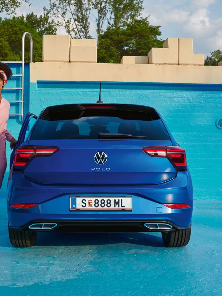 Ein blauer VW Polo steht in einem blau angemalten, leeren Poolbecken, eine junge Frau steigt ein.