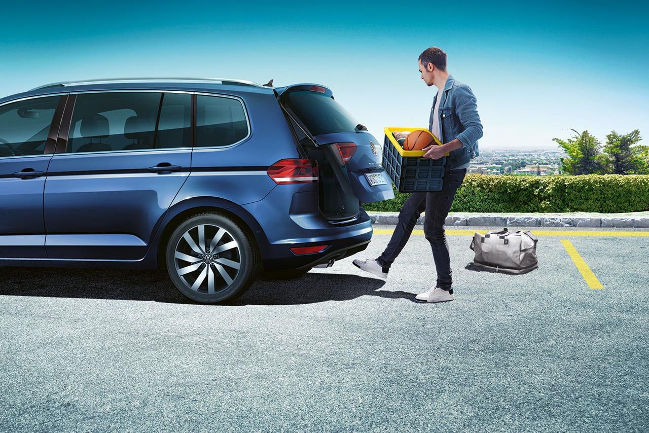 Mann mit Kiste in beiden Händen öffnet Heckklappe eines blauen VW Touran durch Bewegung mit dem Fuß dank  optionaler „Easy Open“ und „Easy Close“-Funktion.