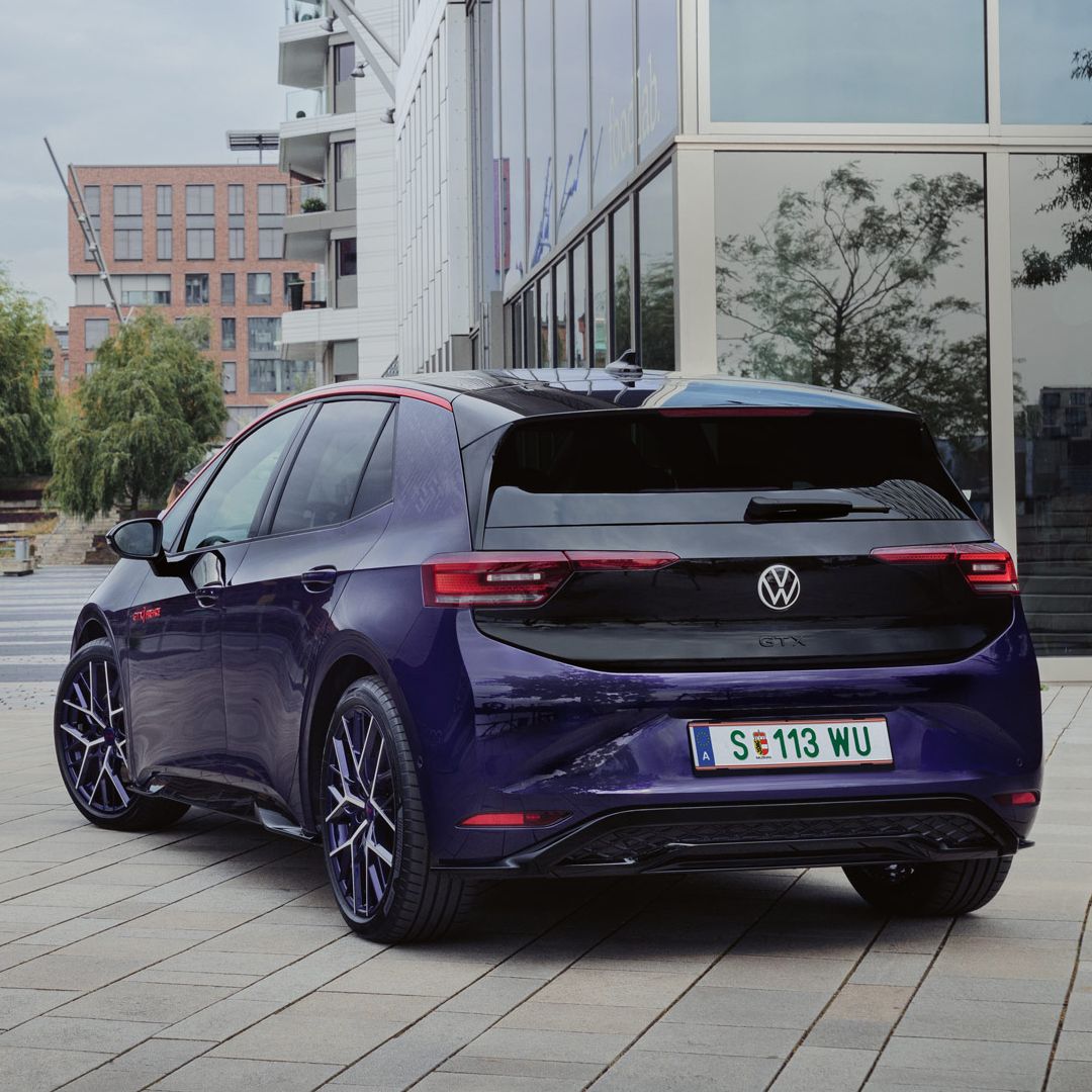 Elektrisches Volkswagen Auto geparkt auf einem modernen Stadtplatz. Im Hintergrund sind Gebäude und eine Brücke zu sehen. Das Auto hat ein Kennzeichen aus Wolfsburg.