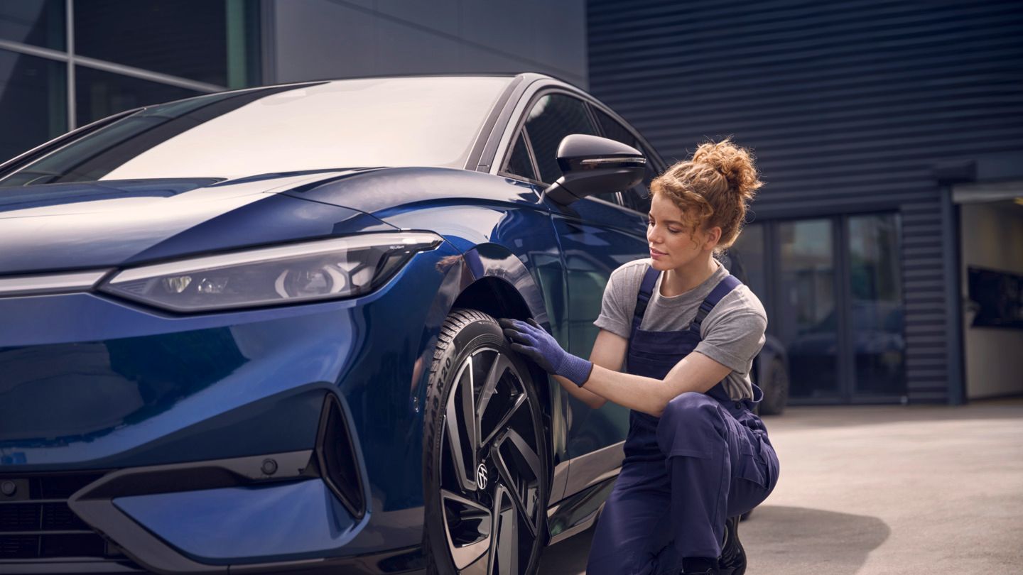 Eine Frau im blauen Overall hockt neben einem VW ID.7 und prüft den Vorderreifen.