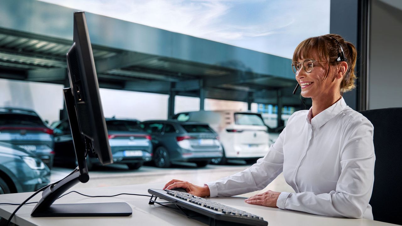 Eine Frau mit weißer Bluse arbeitet an einem Schreibtisch mit einem Computer, headsettragend. Im Hintergrund sind mehrere geparkte Autos und ein überdachter Parkplatz sichtbar.