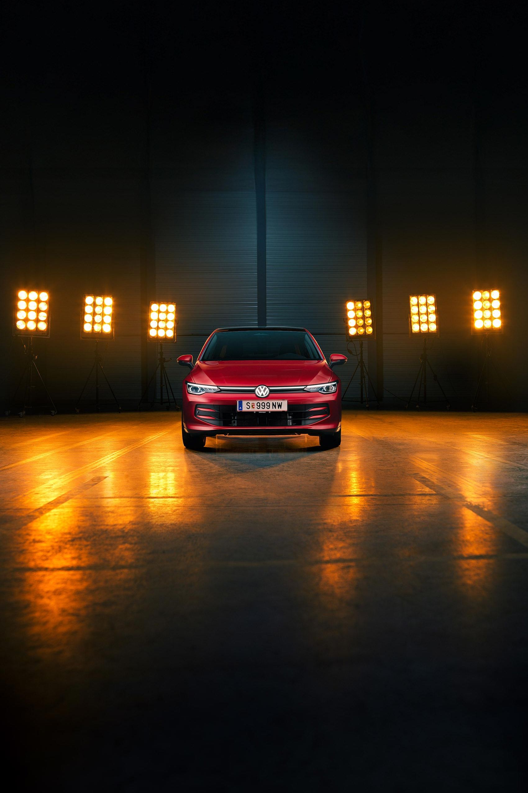 Ein roter VW Golf Falco Edition in Frontalansicht steht mittig im Bild in einer leeren Halle, Bühnenlichter strahlen das Fahrzeug von hinten an.
