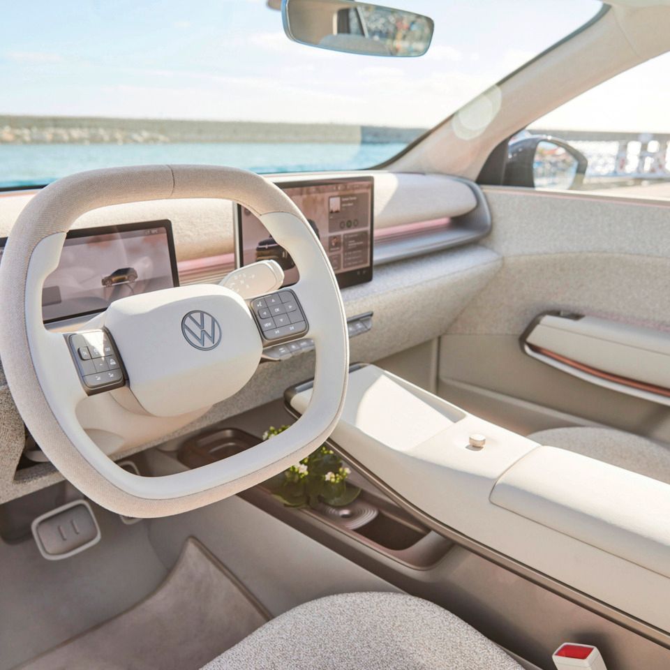 Das Bild zeigt den Innenraum eines ID. Cross mit hellgrauer Polsterung. Das Lenkrad ist zentral platziert und es gibt mehrere digitale Displays auf der Armaturentafel. Im Hintergrund ist ein Wasserblick durch die Frontwindschutzscheibe zu sehen.