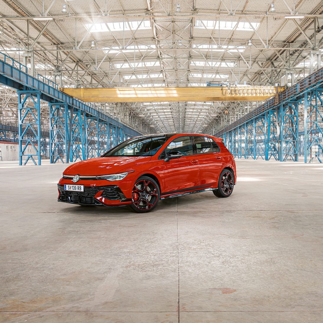 Ein roter Golf GTI steht in einer großen, leeren Industriehalle mit blauen Stahlträgern. Das Dach der Halle ist aus Glas und lässt viel Licht herein.