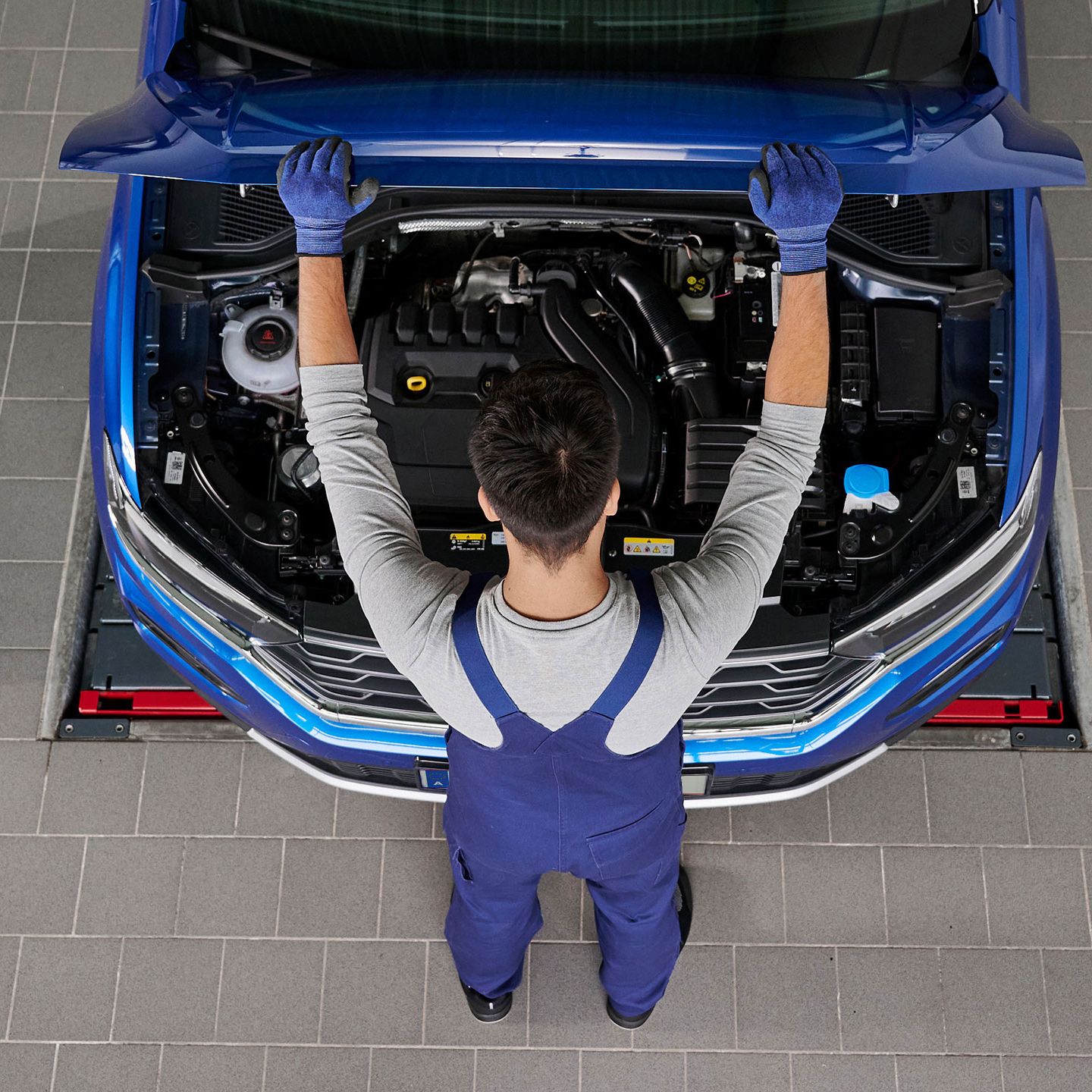 Mechaniker mit blauer Arbeitskleidung arbeitet unter der Motorhaube eines Autos. Der Motorraum ist deutlich sichtbar. Der Mechaniker steht auf grauem Fliesenboden.