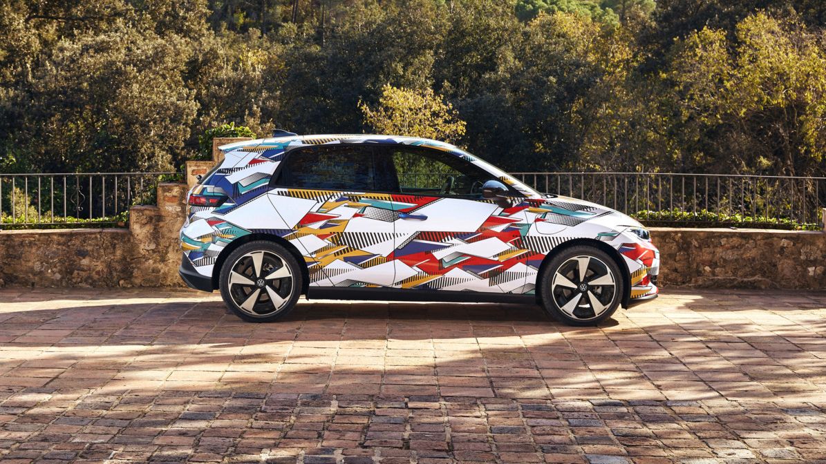 Ein ID. Polo mit buntem, geometrisch gemustertem Aufdruck steht auf einem gepflasterten Platz. Hinter dem Auto erstreckt sich eine bewaldete, hügelige Landschaft. Die Sonne scheint, und das Wetter ist klar.