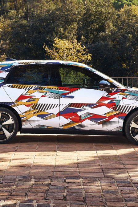 Ein ID. Polo mit buntem, geometrisch gemustertem Aufdruck steht auf einem gepflasterten Platz. Hinter dem Auto erstreckt sich eine bewaldete, hügelige Landschaft. Die Sonne scheint, und das Wetter ist klar.