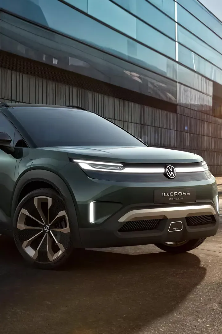 Ein dunkelgrünes Volkswagen ID. Cross steht auf einem Parkplatz vor einem modernen Gebäude mit Glasfassade. Der SUV hat futuristische Scheinwerfer und stylische Felgen. Im Hintergrund sind Treppenstufen und ein paar Palmen sichtbar.