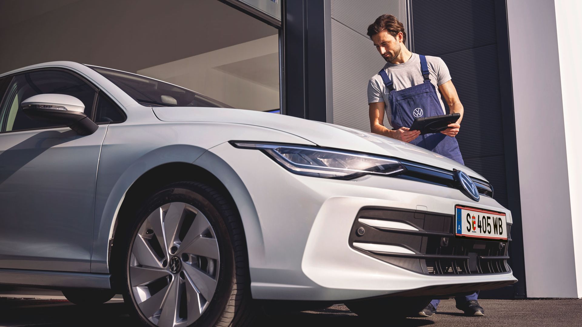 Ein Mechaniker in VW-Arbeitskleidung steht neben einem weißen Golf und hält ein Tablet. 