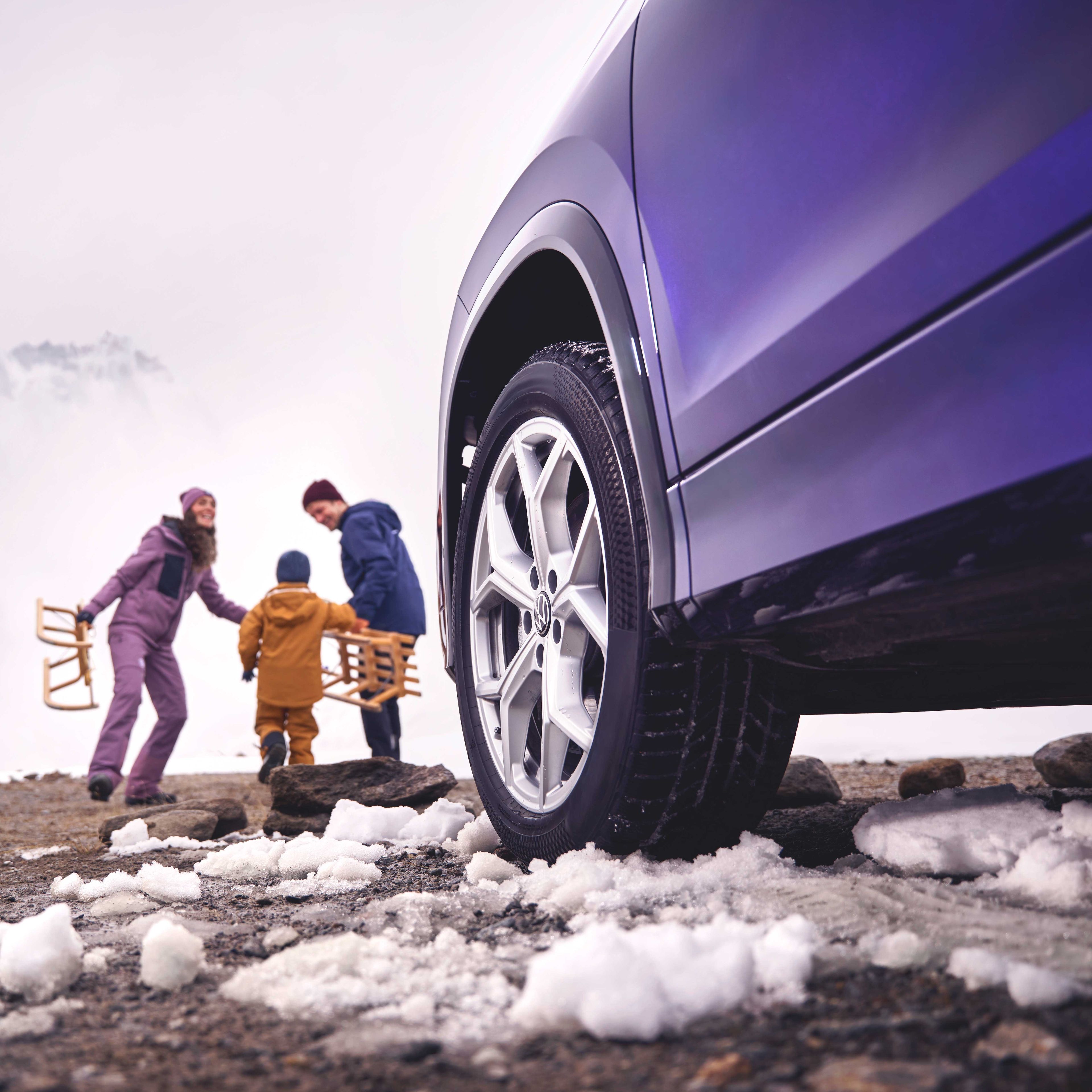 Blick vorbei an der linken Frontseite eines Volkswagen Tayron auf eine Familie mit Schlitten in einer verschneiten Berglandschaft.