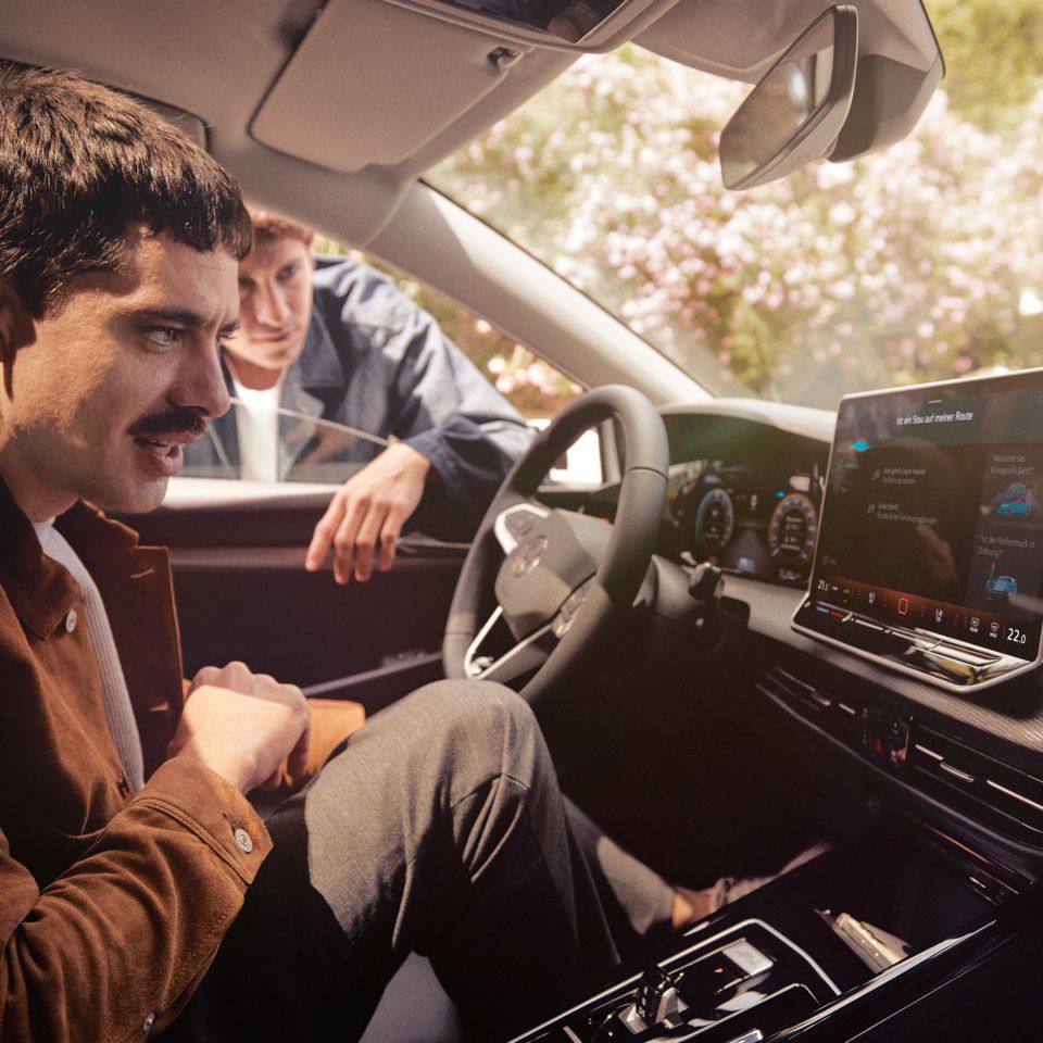 Ein Mann sitzt im neuen VW Golf und schaut auf das Infotainment-Display, ein anderer schaut durch das Fenster.
