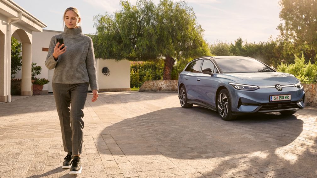 Eine Frau schaut auf ihr Smartphone, im Hintergrund parkt ein blauer VW ID.7 Tourer vor einer Wallbox.