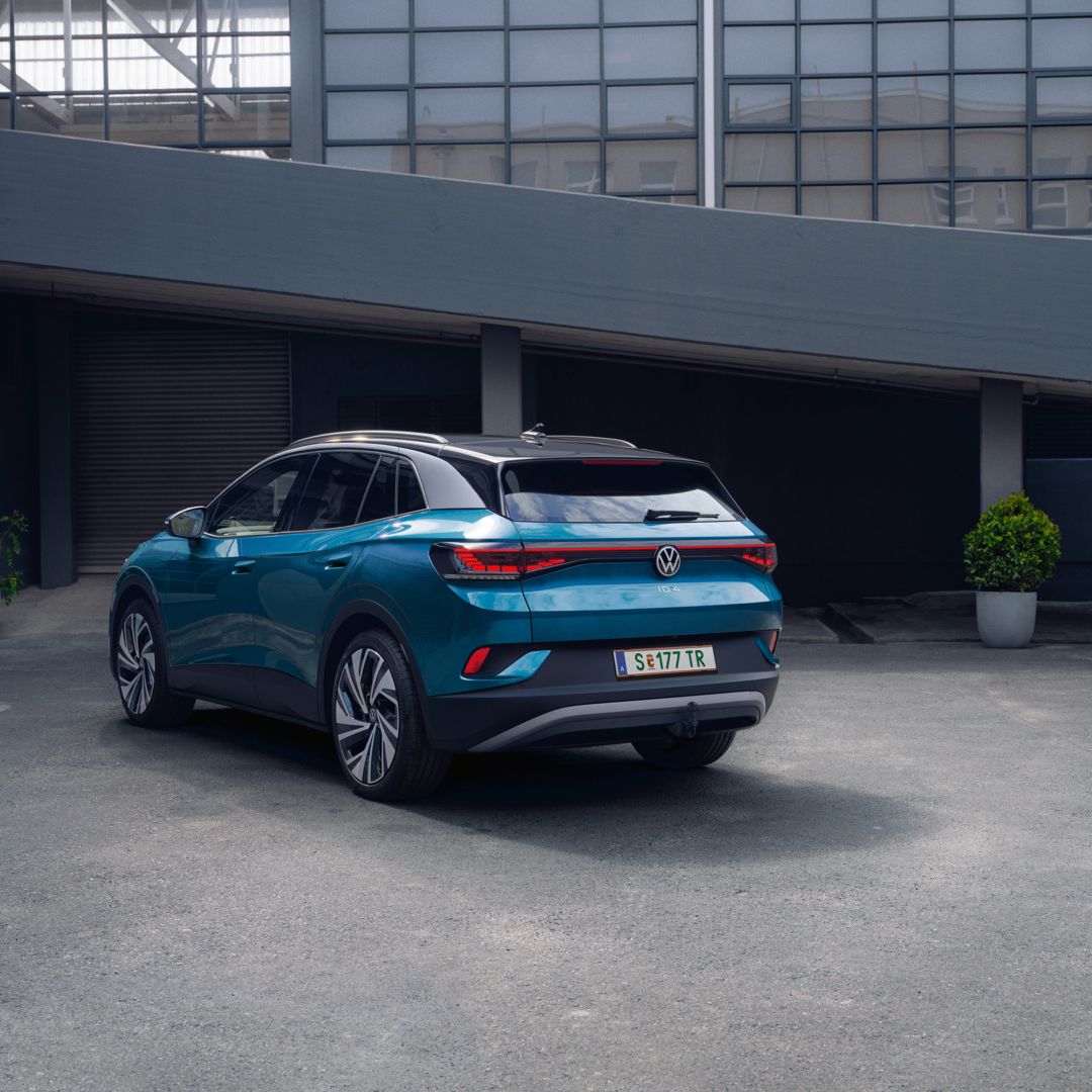 Ein blauer Volkswagen ID.4 steht geparkt in einem modernen, städtischen Umfeld. Das Fahrzeug zeigt seine Rückansicht. Im Hintergrund sind Gebäude aus Glas und Beton zu sehen.
